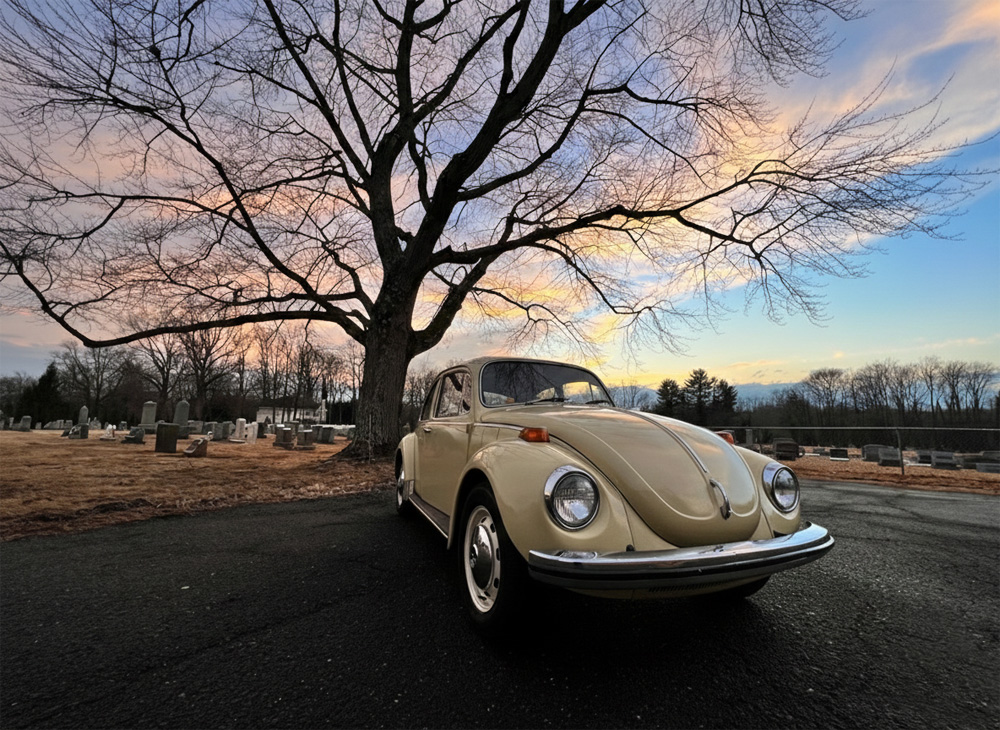 Classic beige Volkswagen Beetle parked under a large tree at sunset with colorful sky and cemetery in the background.