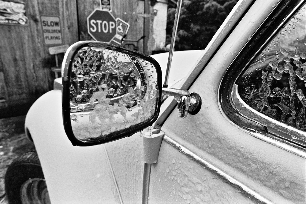 Black and white close-up of vintage Volkswagen Beetle side mirror covered in ice with frosted window and stop sign in background.