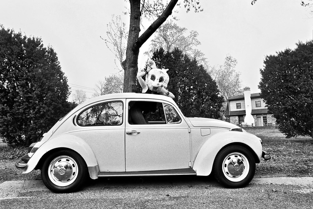 A black and white shot of a 1302 Super Beetle with a girl with a rabbit head on peeking out of the sunroof.