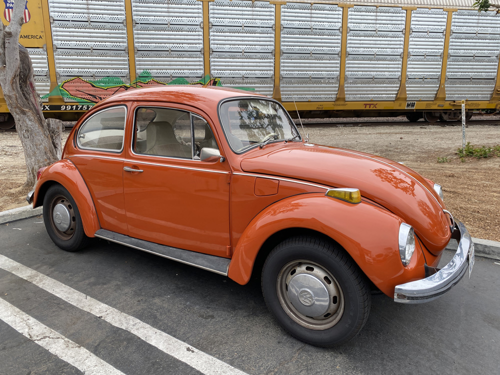 Orange Volkswagen Beetle parked near a freight train with metal panels.