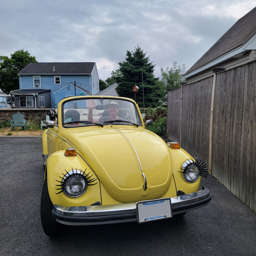 Ellen waving from the driver's seat of her yellow Volkswagen Super Beetle convertible with eyelash headlight accents in Massachusetts