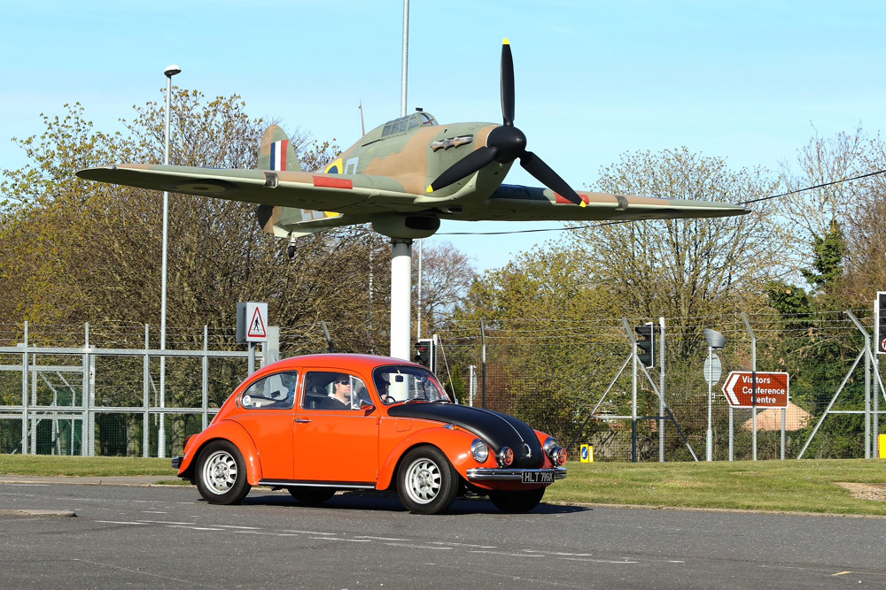 Orange VW Super Beetle parked beneath WWII Hawker Hurricane aircraft on pedestal near visitor center