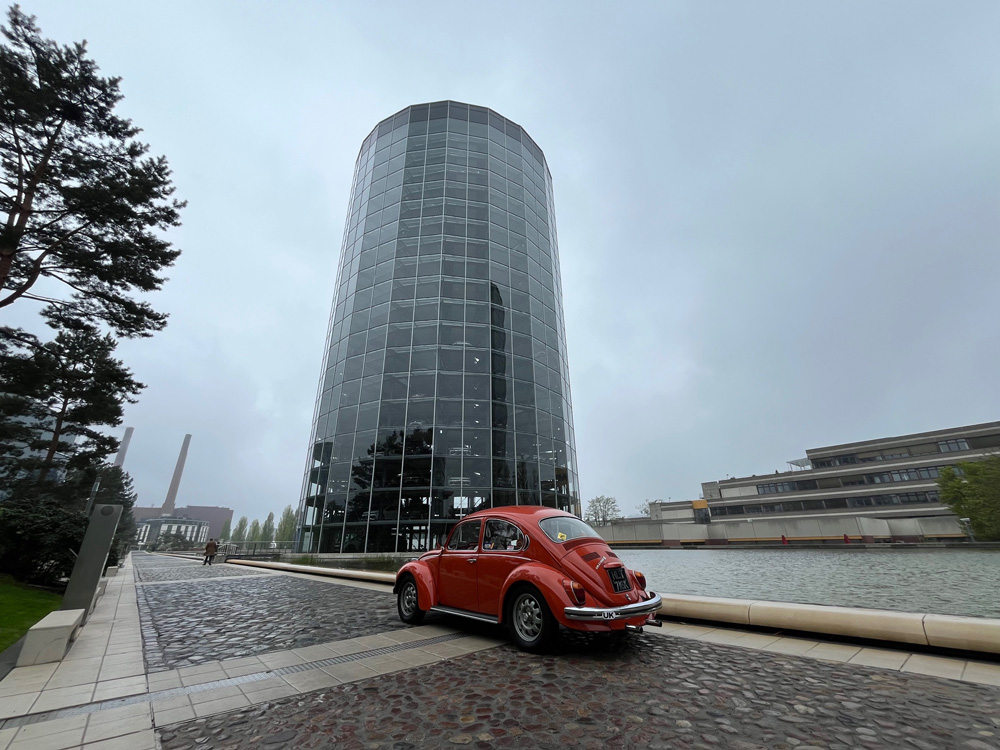 Orange VW Super Beetle parked beside tall glass building near canal under cloudy sky