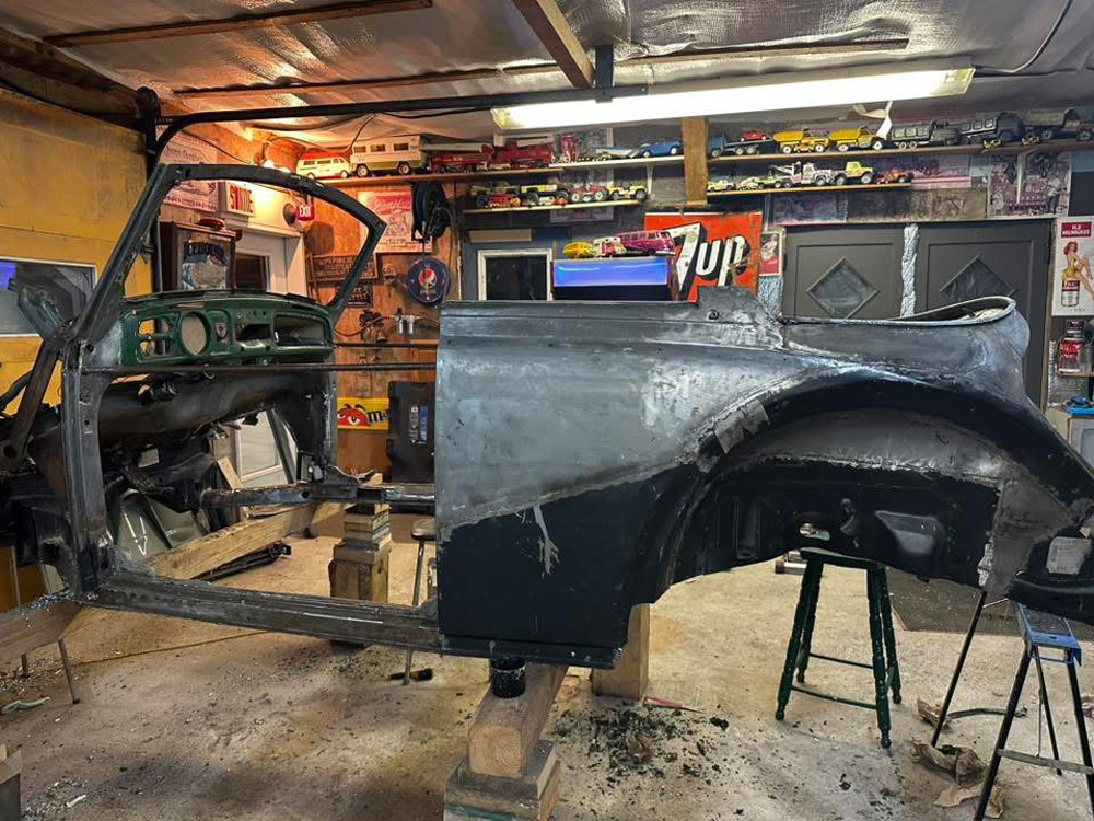 Chopped VW Super Beetle convertible hot rod project in garage: stripped bare-metal body in primer, green dash exposed, on frame stands amid collectible toys and posters.