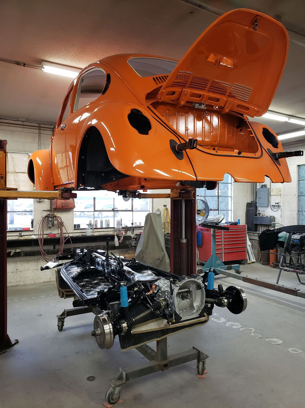 Orange VW Super Beetle convertible body in primer/gloss on hoist over rolling chassis with engine and drivetrain exposed in garage workshop.