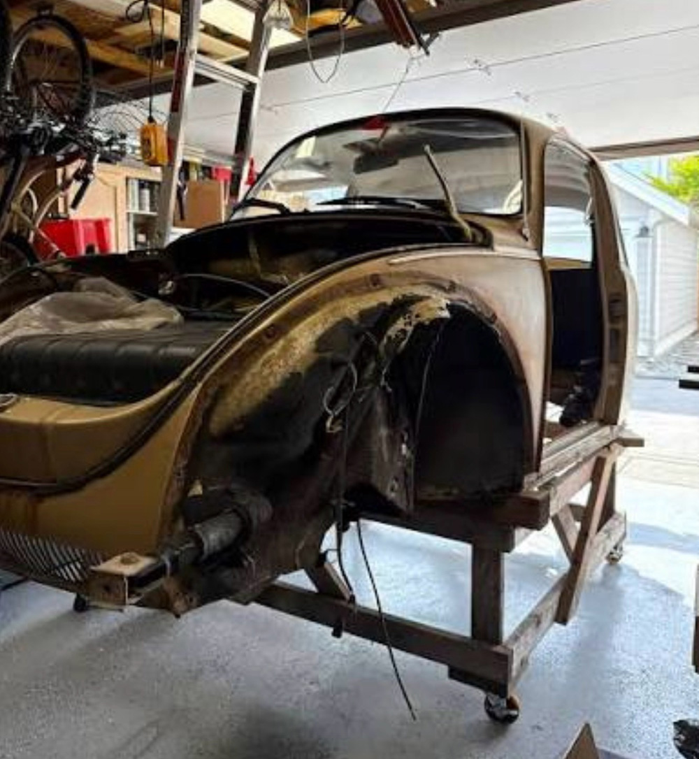 Rusty and patched tan/beige VW Super Beetle body shell in garage, mounted on wheeled dolly stand with open doors and hood, showing interior black seats, exposed metalwork, and workshop clutter including bikes and tools in background.