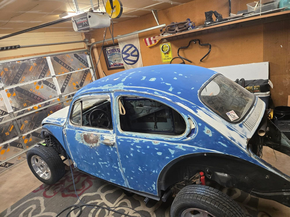 Blue VW Super Beetle coupe in garage, heavily modified with lifted suspension, large off-road tires, flared fenders, patchy blue primer paint, doors off showing stripped interior, mounted on floor amid pegboard walls, VW signs, tools, and workshop clutter.