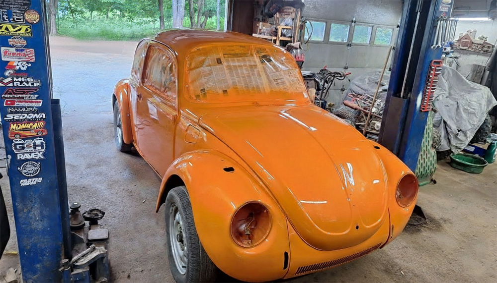 Bright orange VW Super Beetle coupe in garage workshop on lift, freshly painted glossy body with hood closed, front fenders and doors in place, surrounded by sticker-covered walls, tools, and outdoor view of trees and parked cars through open door.