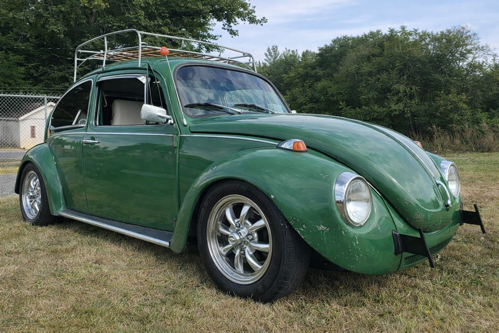 alt="Green Volkswagen Super Beetle with roof rack parked on grass near trees and a fence