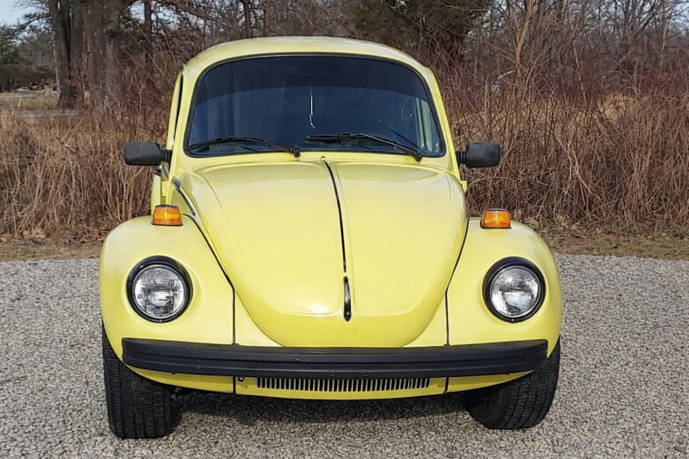 Yellow Volkswagen Super Beetle parked on gravel with trees in the background