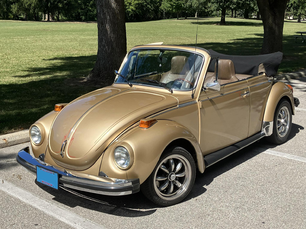 Metallic gold Volkswagen Super Beetle convertible parked near grass and trees.