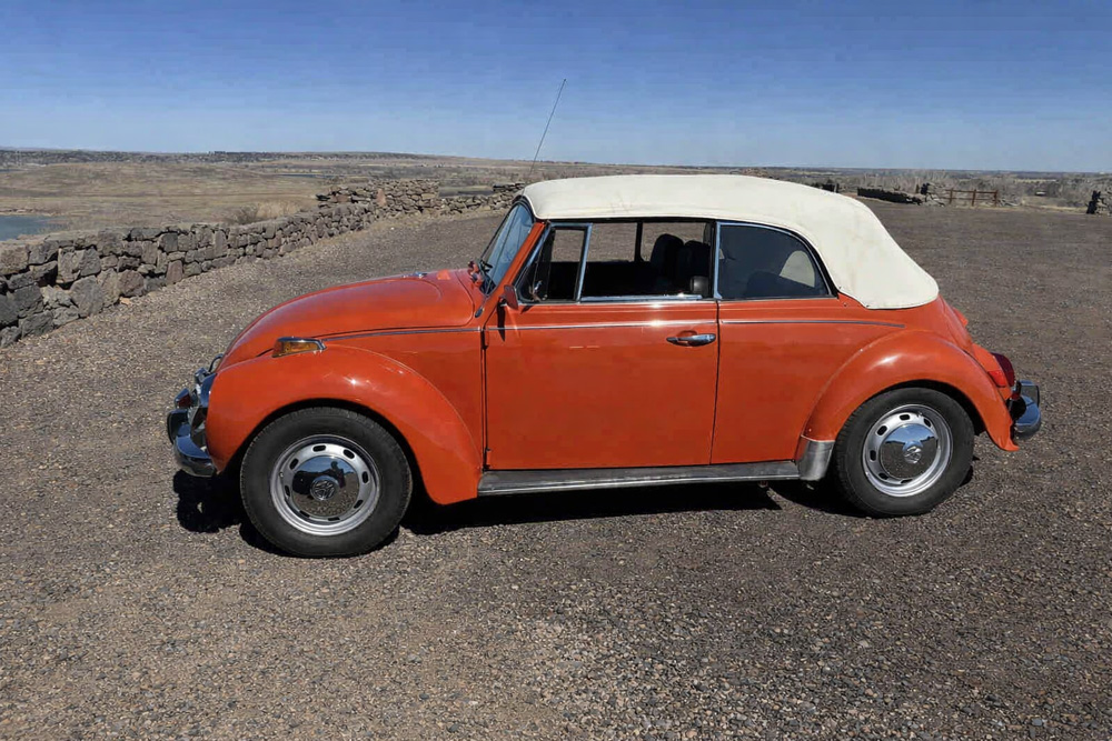 Orange Volkswagen Super Beetle convertible parked on gravel near a stone wall.