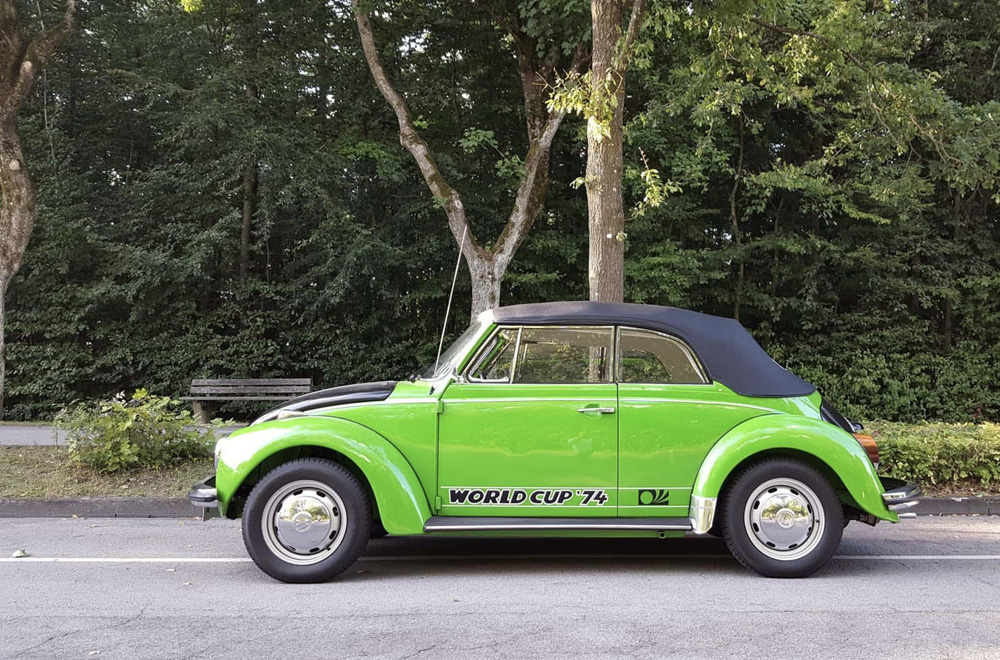 Side profile of a restored green 1974 World Cup Super Beetle convertible with black top and World Cup '74 graphics parked along a tree-lined road