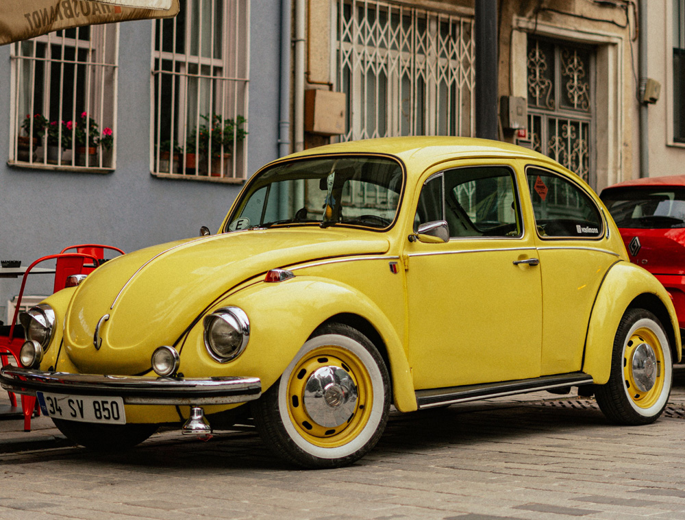 Yellow Volkswagen Super Beetle parked on a cobblestone street with flower boxes in the background