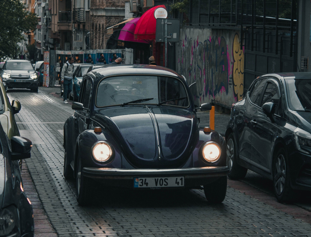 Blue Volkswagen Super Beetle driving down a narrow cobblestone street in a busy urban area