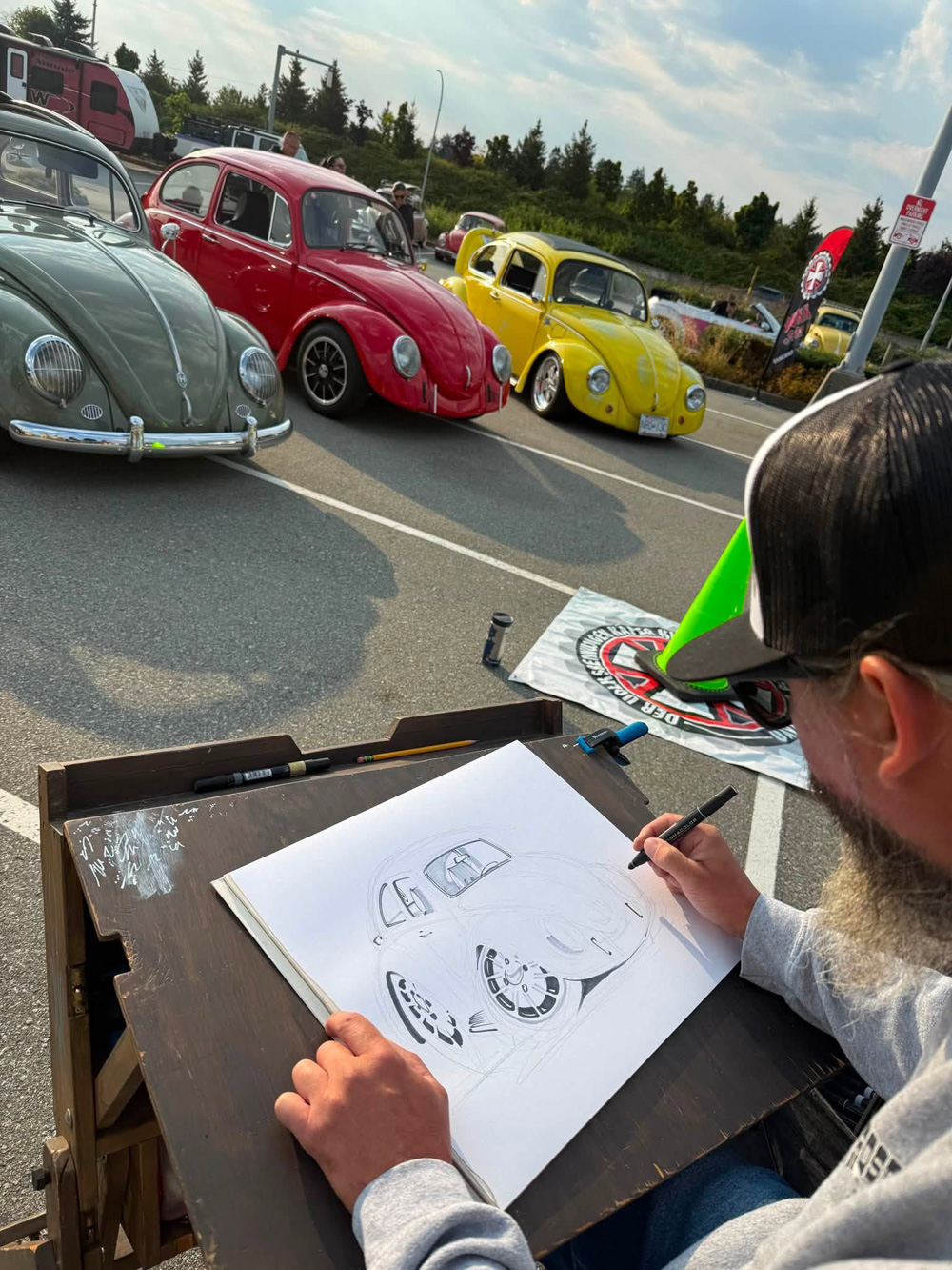 Artist drawing a Volkswagen Beetle at an outdoor meet with several Beetles parked behind the sketch