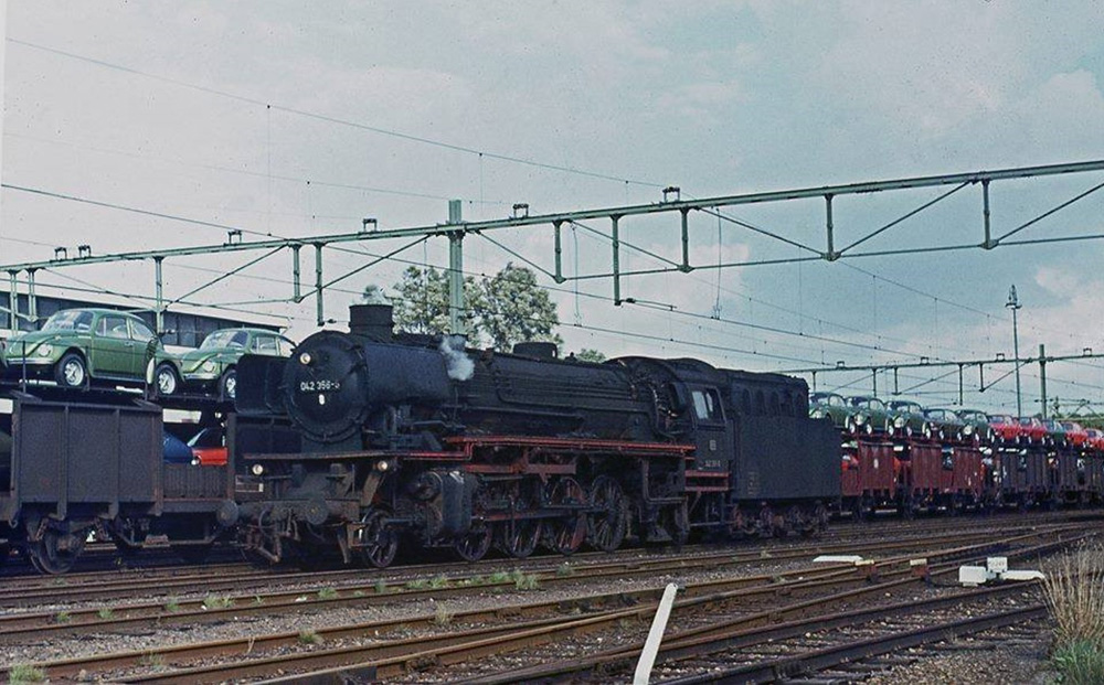 Deutche Bundesbahn Class 42 steam locomotive 042 356 stands next to a 'Pon train' loaded with new Volkswagens